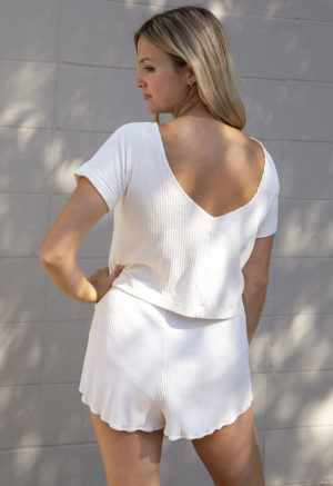 Back view of a straight size model in Ivory Rib Short Shorts, standing outside in front of white brick wall.