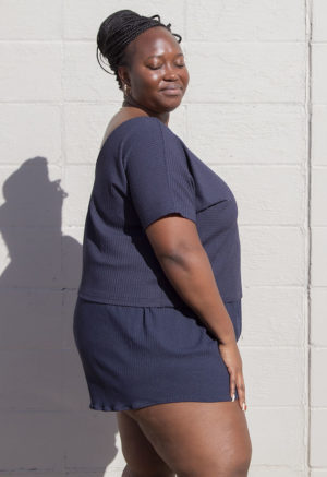 Side view of plus size model in Navy Rib Reversible Short Sleeve Top standing in front of white brick wall.