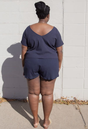 Back view of plus size model in Navy Rib Reversible Short Sleeve Top standing in front of white brick wall.
