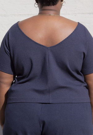 Back view of plus size model in Navy Rib Reversible Short Sleeve Top standing in front of white brick wall.