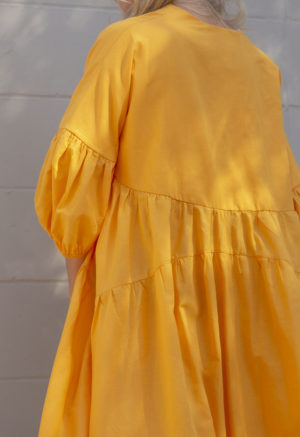 Back view of straight size model in Gold Circle Midi Dress standing in front of white brick wall.