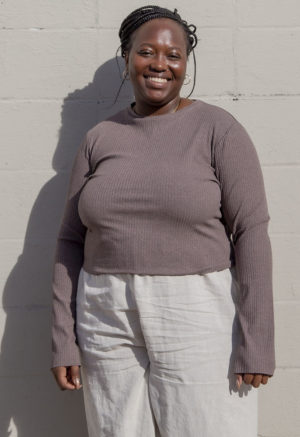 Front view of a plus size model in the Mauve Rib Cropped Long Sleeve Top, standing in front of white brick wall.