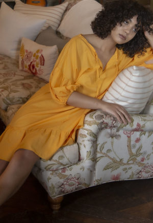 Front view of straight size model in Gold Reversible Ruffle Dress sitting on couch in French-inspired apartment.