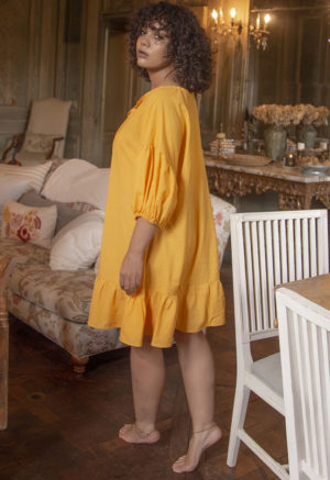 Back/side view of straight size model in Gold Reversible Ruffle Dress standing in French-inspired apartment.