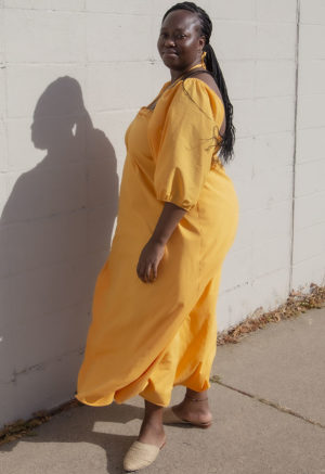 Side view of plus size model in Gold Keyhole Maxi Dress standing in front of white brick wall.