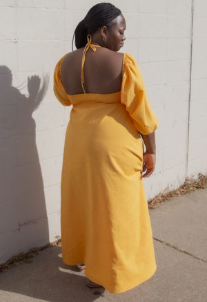 Back view of plus size model in Gold Keyhole Maxi Dress standing in front of white brick wall.