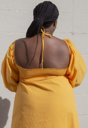 Close-up back view of plus size model in Gold Keyhole Maxi Dress standing in front of white brick wall.