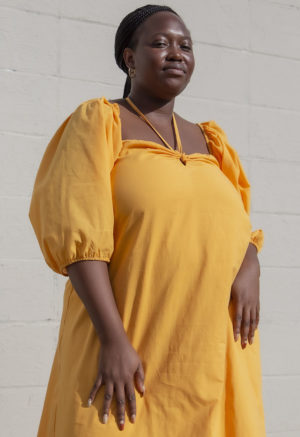 Front view of plus size model in Gold Keyhole Maxi Dress standing in front of white brick wall.