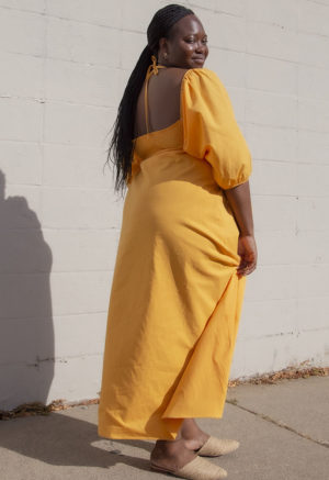 Back/side view of plus size model in Gold Keyhole Maxi Dress standing in front of white brick wall.