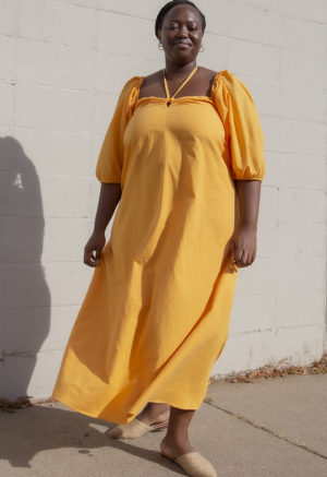 Front view of plus size model in Gold Keyhole Maxi Dress standing in front of white brick wall.
