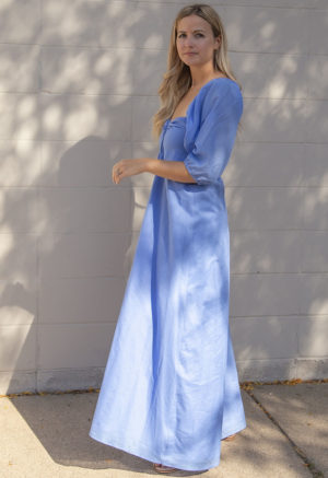 Side view of straight size model in Cerulean Blue Keyhole Maxi Dress standing in front of white brick wall.