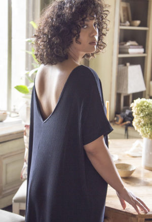 Back view of a straight size model in the Navy Rib Reversible Side Slit Dress, standing by wooden table.