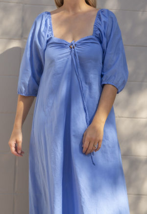Close-up front view of straight size model in Cerulean Blue Keyhole Maxi Dress standing in front of white brick wall.