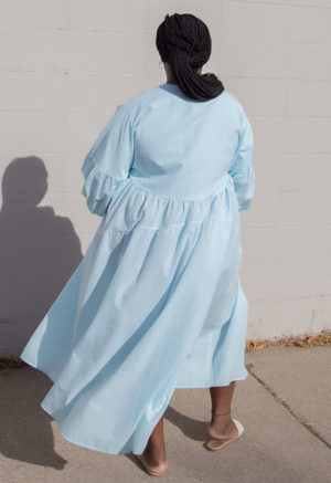 Back view of plus size model in Capri Blue Circle Midi Dress standing outside in front of white brick wall.