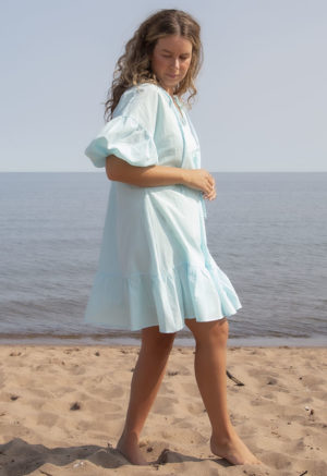 Side view of straight size model in Capri Blue Reversible Ruffle Dress standing on beach with lake in background.
