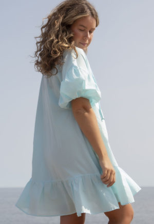 Back/side view of straight size model in Capri Blue Reversible Ruffle Dress standing outside.