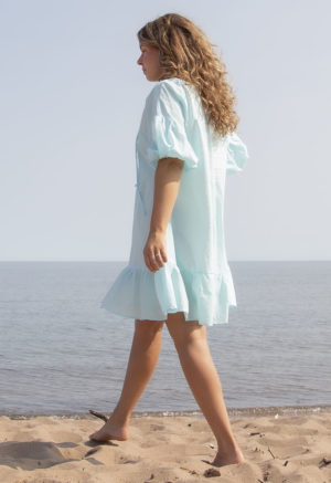 Back/side view of straight size model in Capri Blue Reversible Ruffle Dress standing on beach with lake in background.
