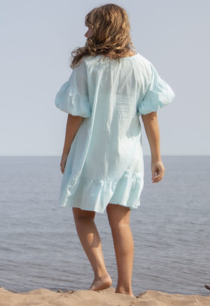 Back view of straight size model in Capri Blue Reversible Ruffle Dress standing on beach with lake in background.