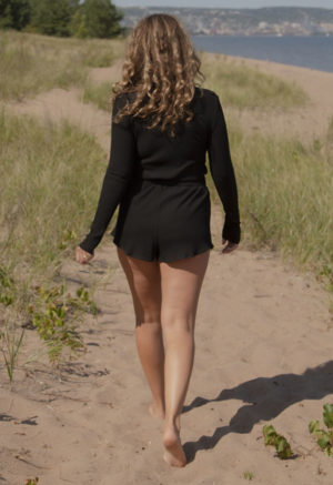 Back view of a straight size model in the Black Rib Cropped Long Sleeve Top, walking outside on beach path.