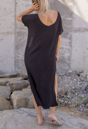 Back view of a straight size model in the Black Rib Reversible Side Slit Dress, standing in front of concrete wall.