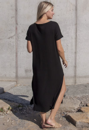 Back view of a straight size model in the Black Rib Reversible Side Slit Dress, standing in front of concrete wall.