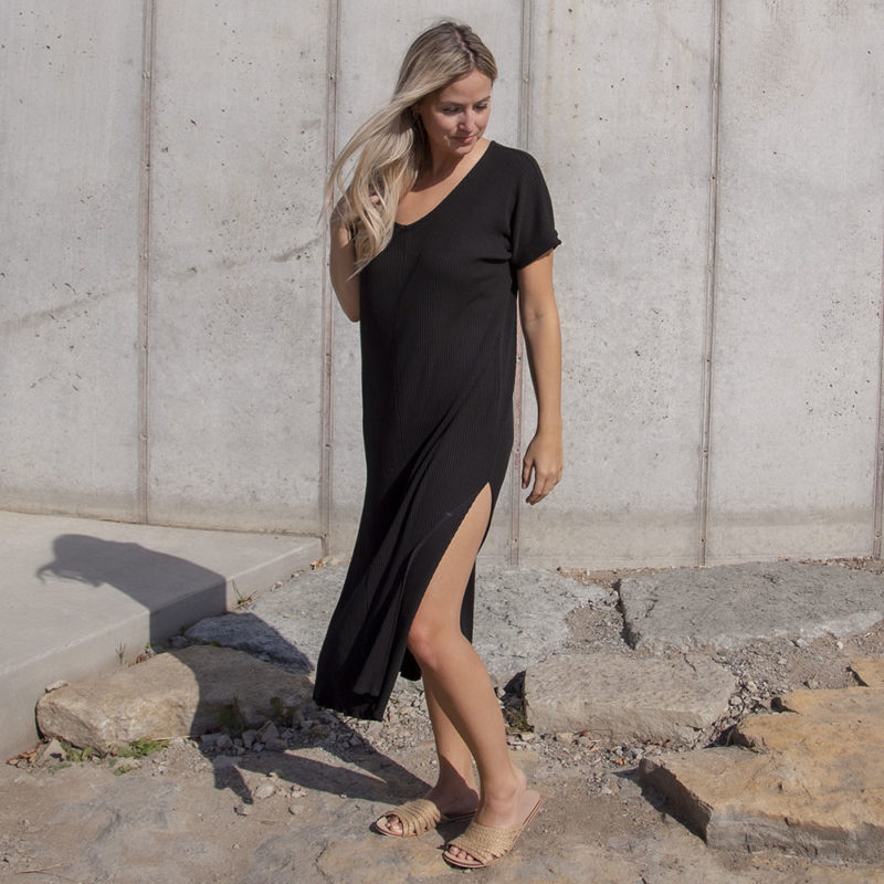 Front/side view of a straight size model in the Black Rib Reversible Side Slit Dress, standing in front of concrete wall.