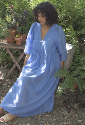 Front/side view of straight size model in Cerulean Blue Balloon Sleeve Midi Dress sitting outside in front of bushes.