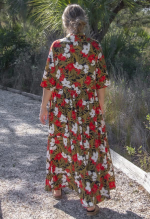 Back view of straight size model wearing Island Floral Tiered Lapel Dress standing on path with bushes behind her.