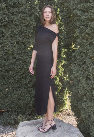 Woman in black rib maxi length dress with an asymmetrical neck line and high slit on one side of the skirt